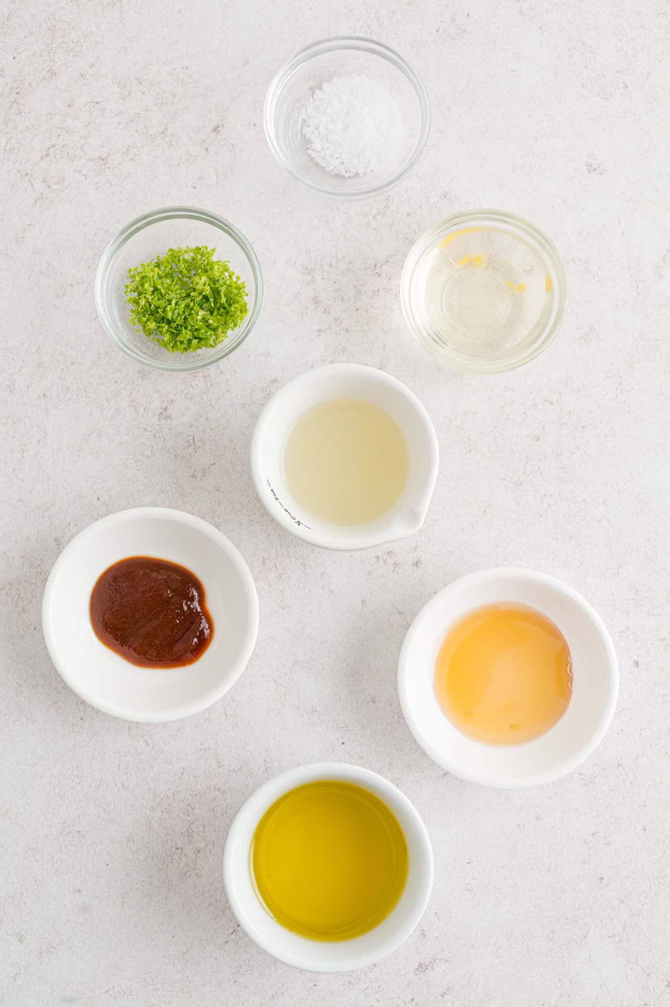 Seven small bowls containing various ingredients, including lime zest, lime juice, olive oil, honey, salt, hot sauce, and a clear liquid, arranged on a light-colored surface.