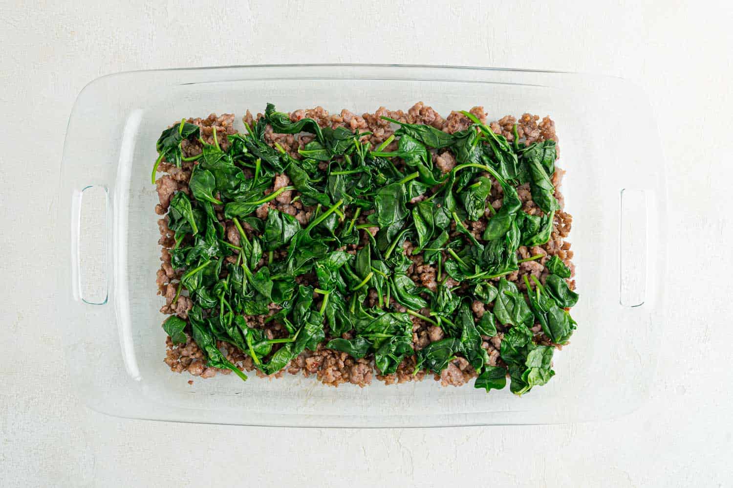 Spinach on top of ground sausage in a baking dish.