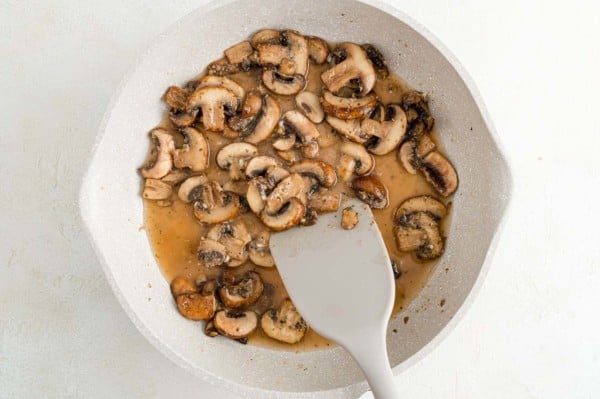 A spatula stirs sauteed mushrooms with marsala wine in a skillet.