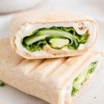 Two halves of a turkey wrap on a white plate, with an avocado and bowl of cucumber slices in the background.