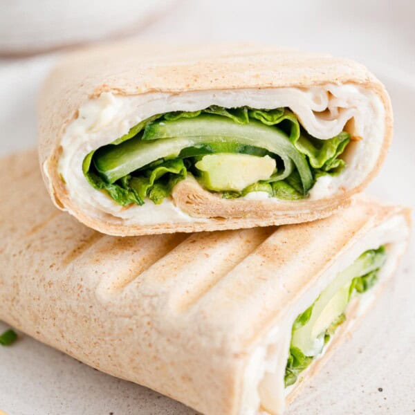 Two halves of a turkey wrap on a white plate, with an avocado and bowl of cucumber slices in the background.