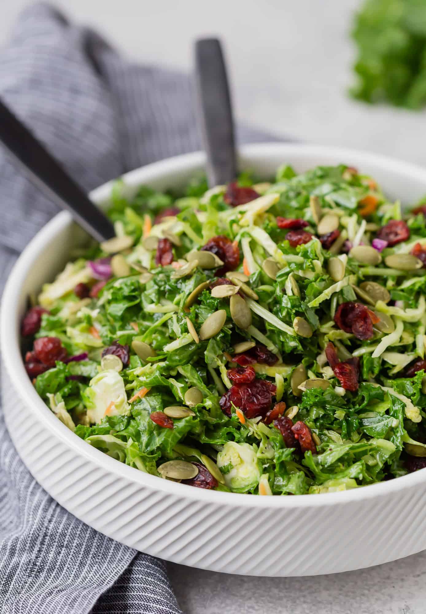 Sweet kale salad in white bowl with black utensils.