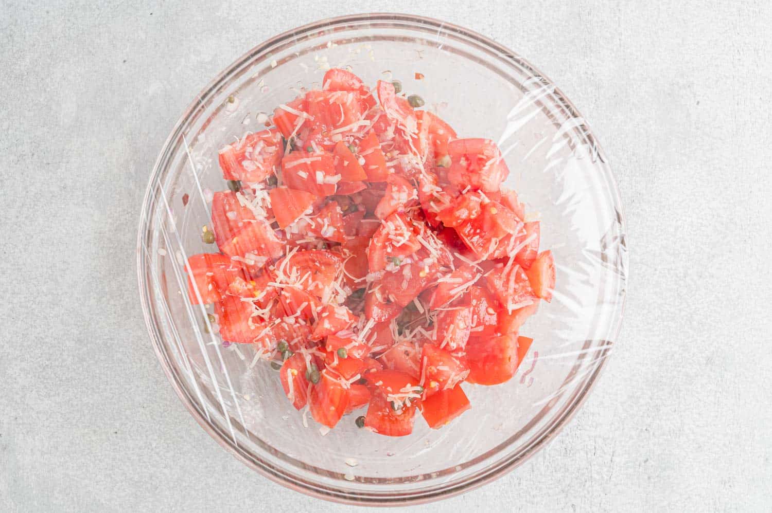 Tomatoes marinating in a glass bowl covered with plastic wrap.