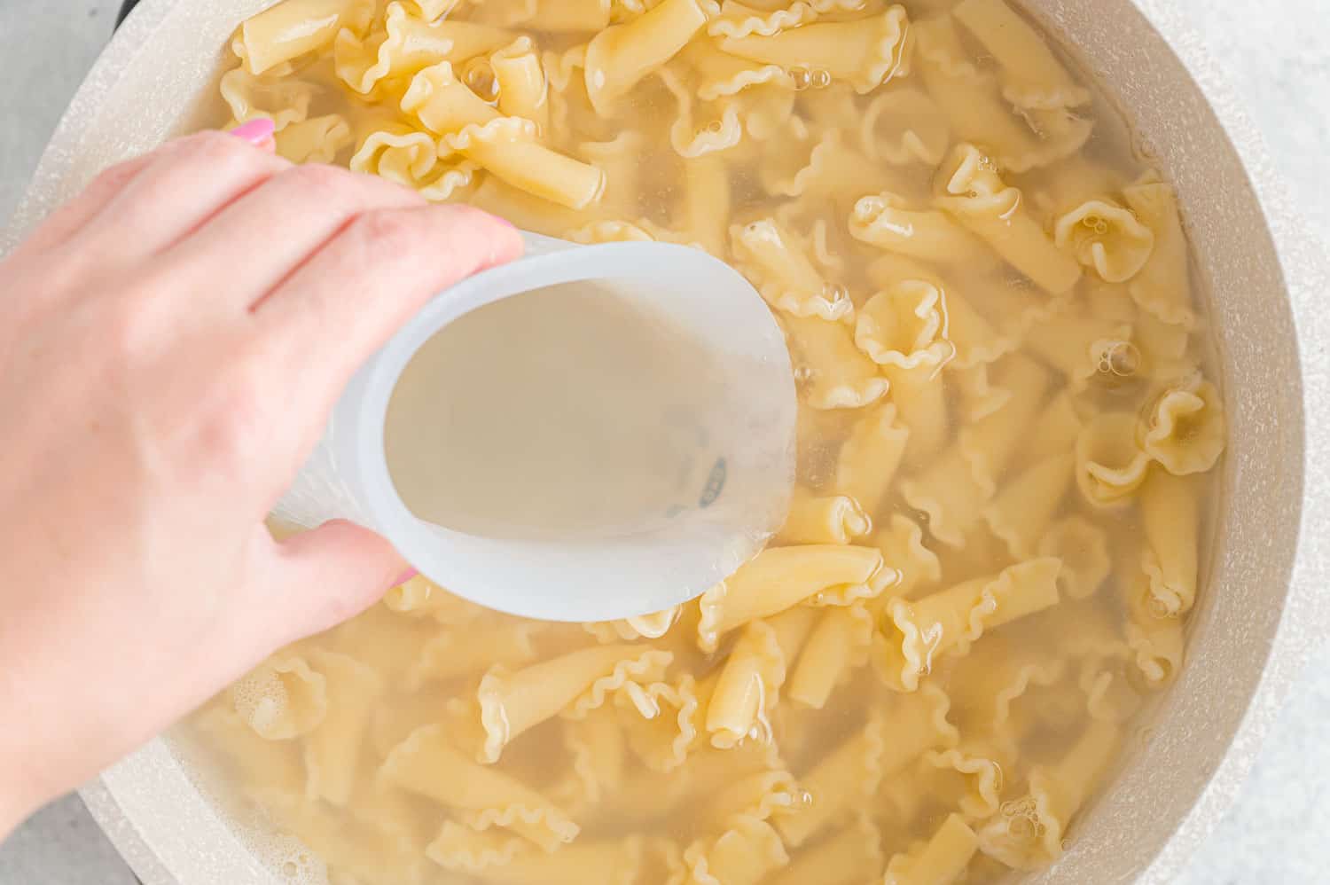 A hand scooping a cup of reserved pasta water from a pot of cooked pasta.