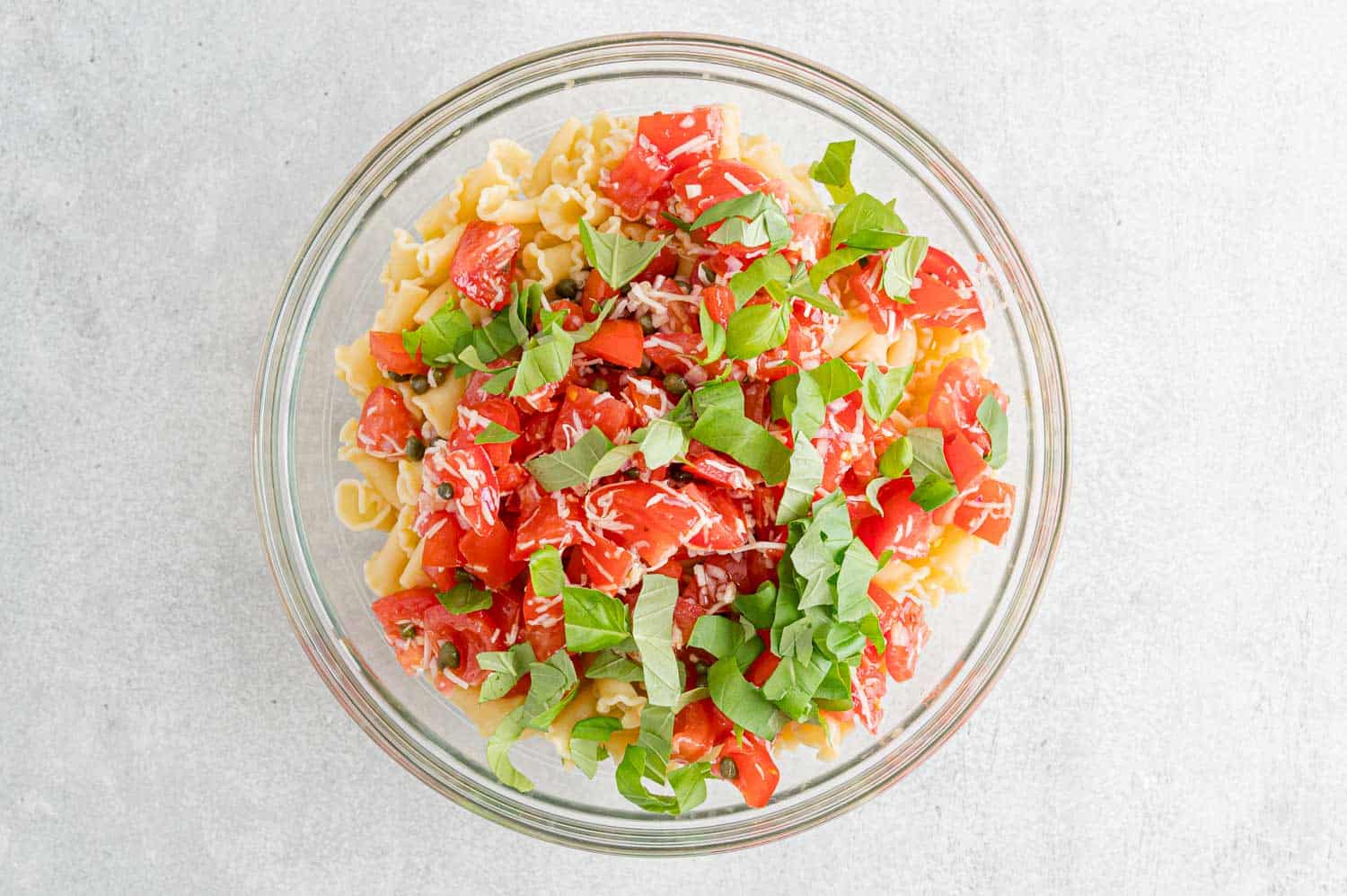 Fresh marinated tomatoes, basil, and seasonings added to a glass bowl with cooked pasta.