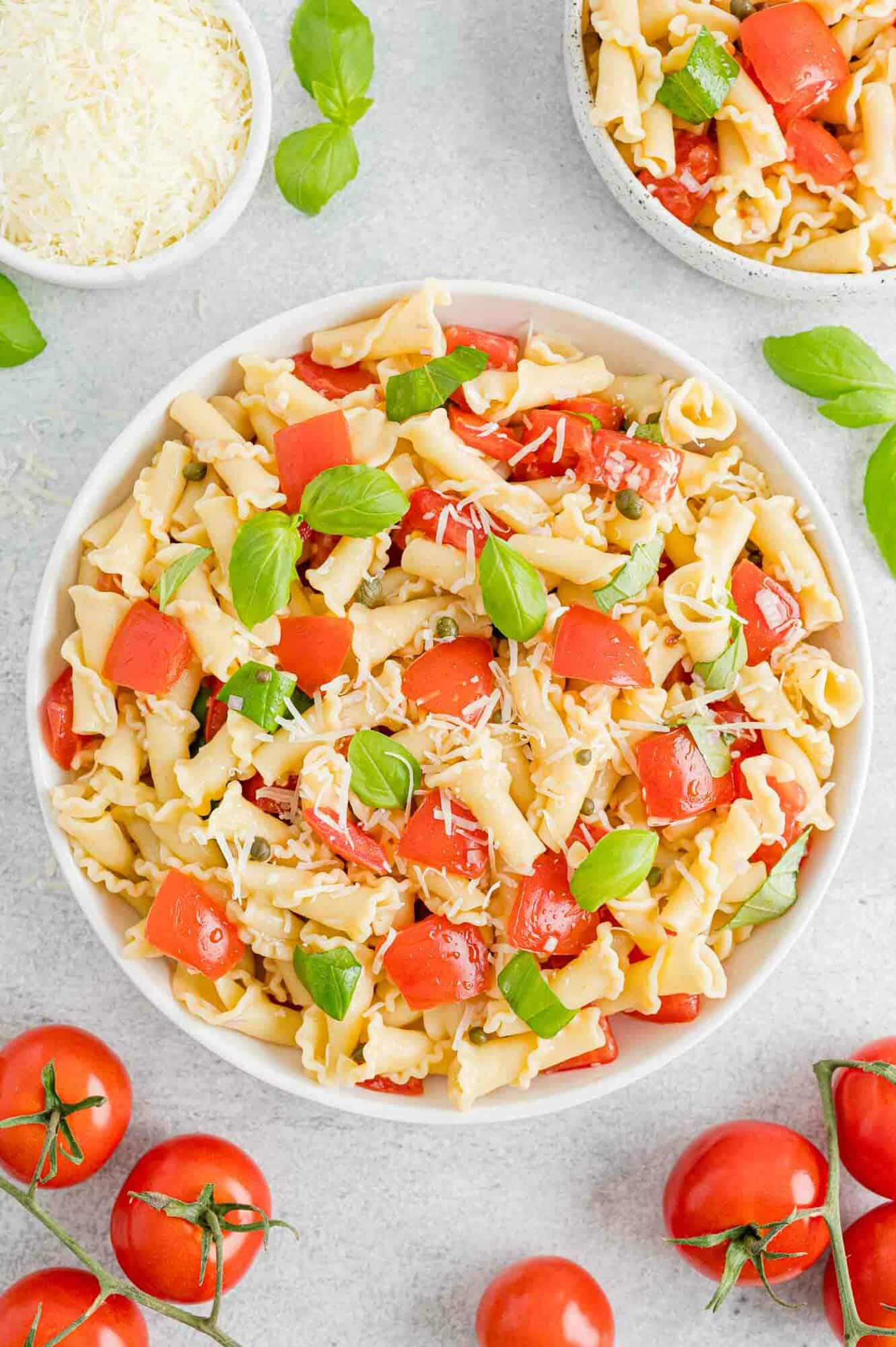 Overhead view of a bowl of fresh tomato pasta surrounded by scattered tomatoes and a bowl of grated parmesan.