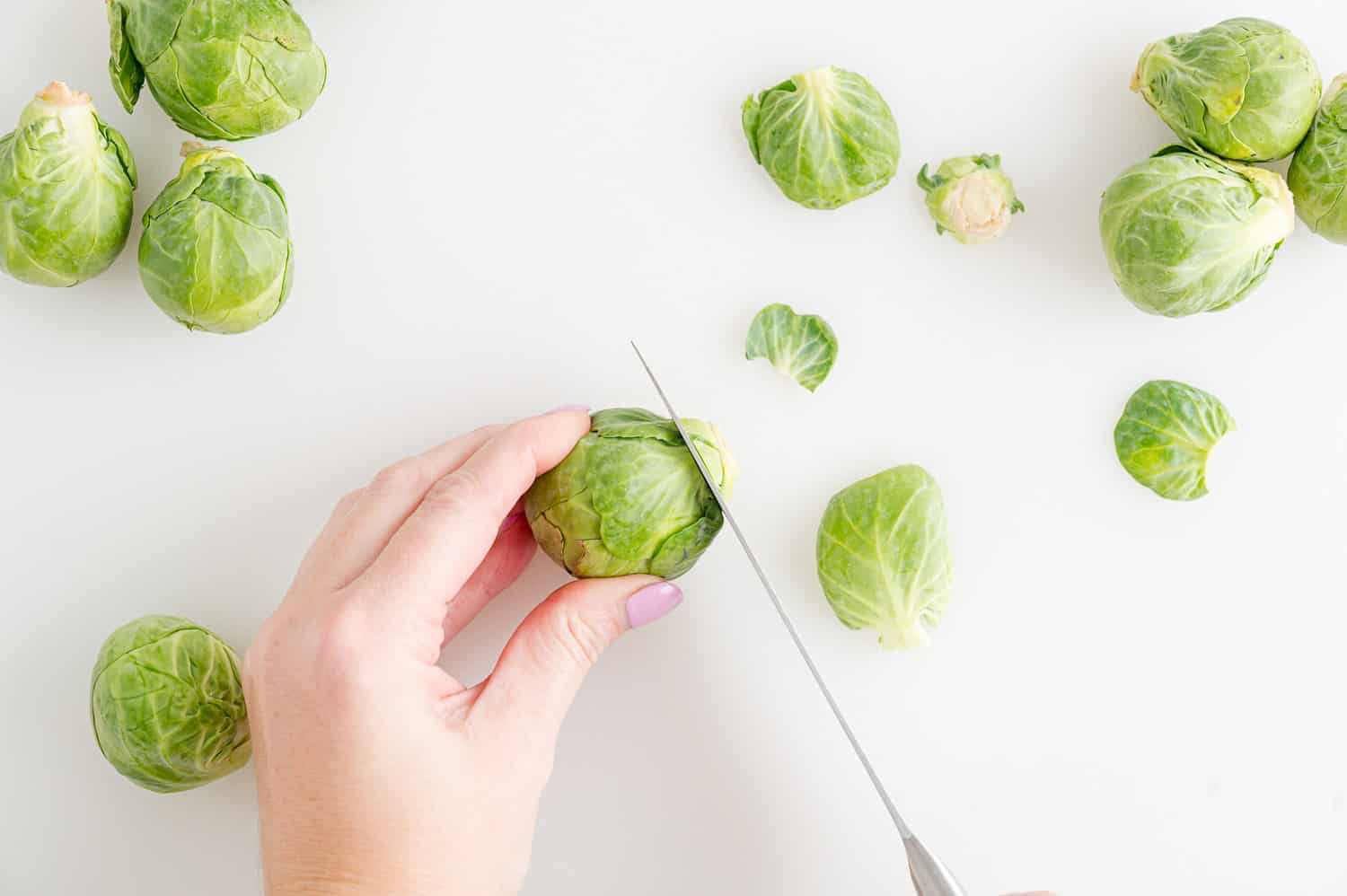 Overhead view of a hand holding a whole Brussels sprout while using a knife to trim off the stem.