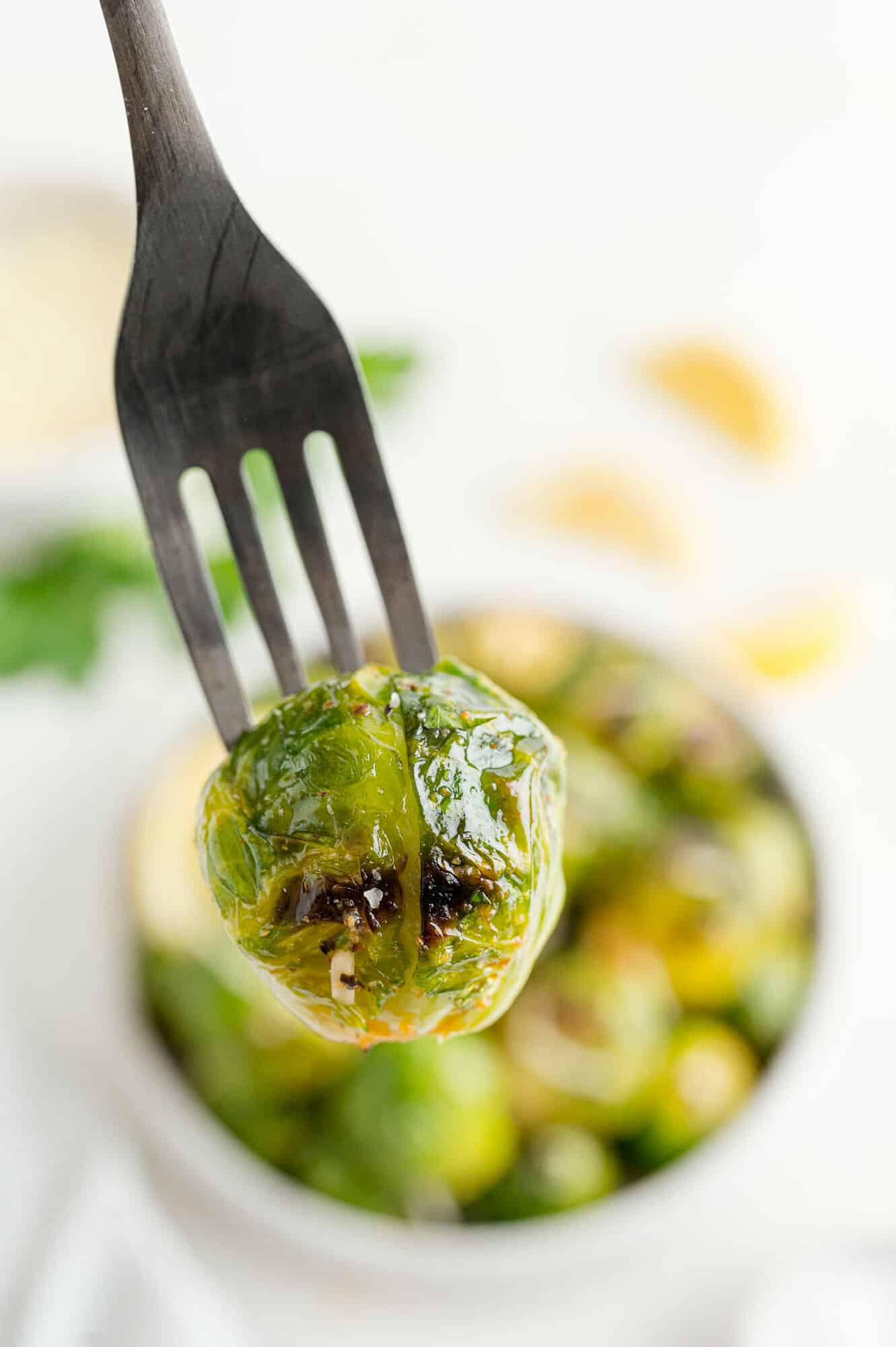 Close up of a grilled Brussels sprout on a fork, held over a bowl of Brussels sprouts in the background.