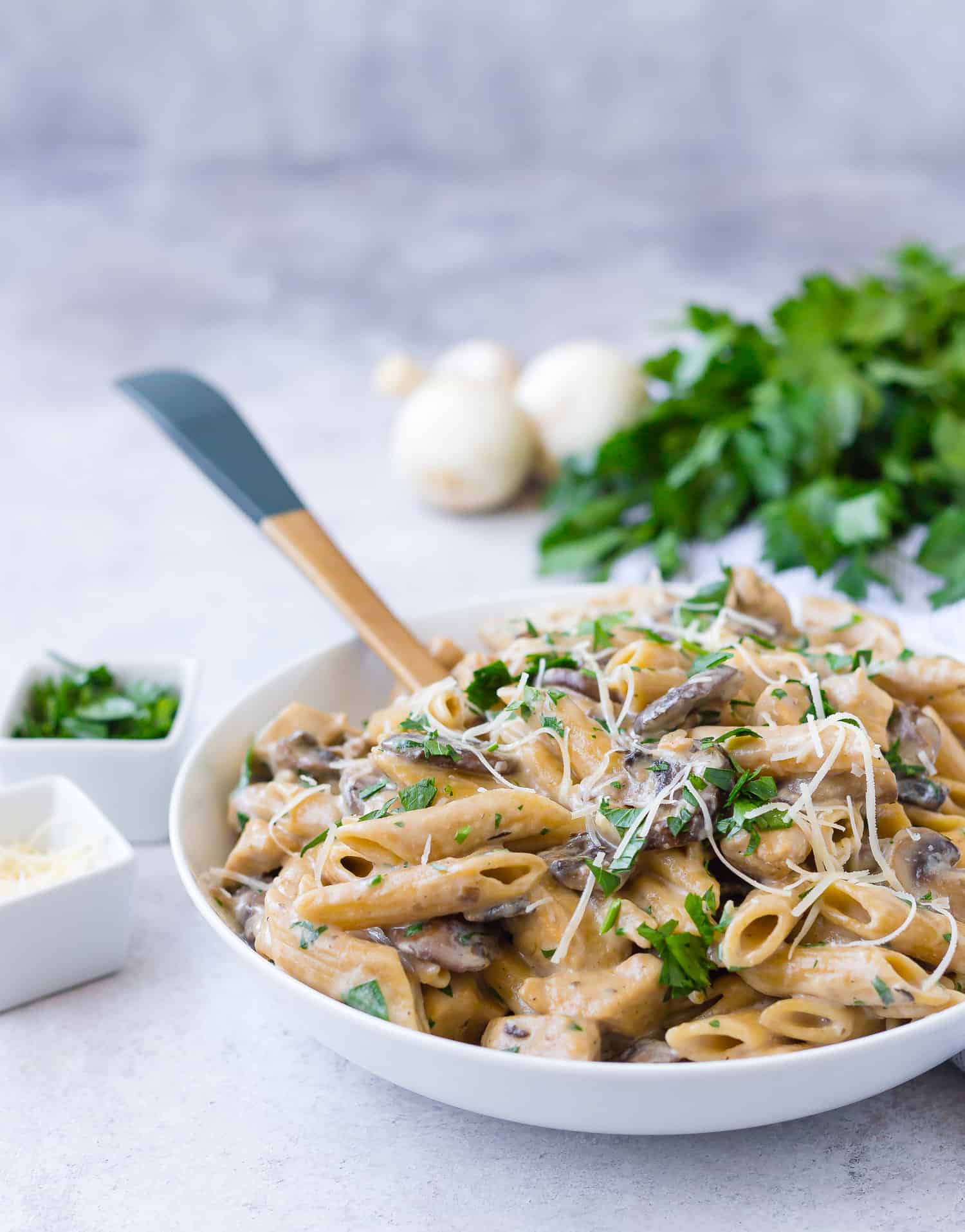 Pasta in serving bowl with mushrooms and parsley in background.