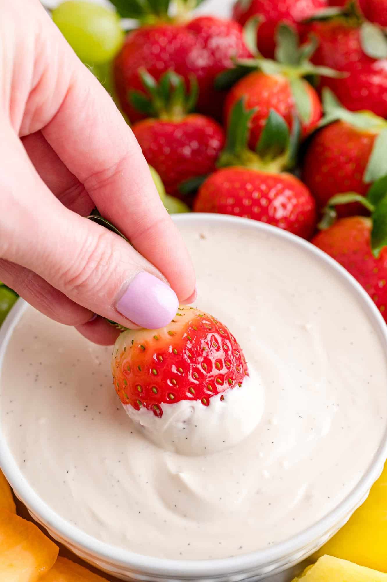 A hand dips a strawberry into a bowl of cream cheese fruit dip in the center of a fruit platter.