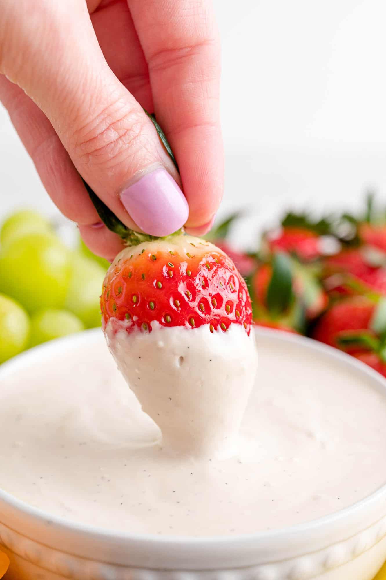 A hand dips a strawberry into a bowl of cream cheese fruit dip in the center of a fruit platter.