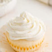 An unwrapped vanilla cupcake topped with a swirl of vanilla buttercream frosting, with a piping bag in the background.