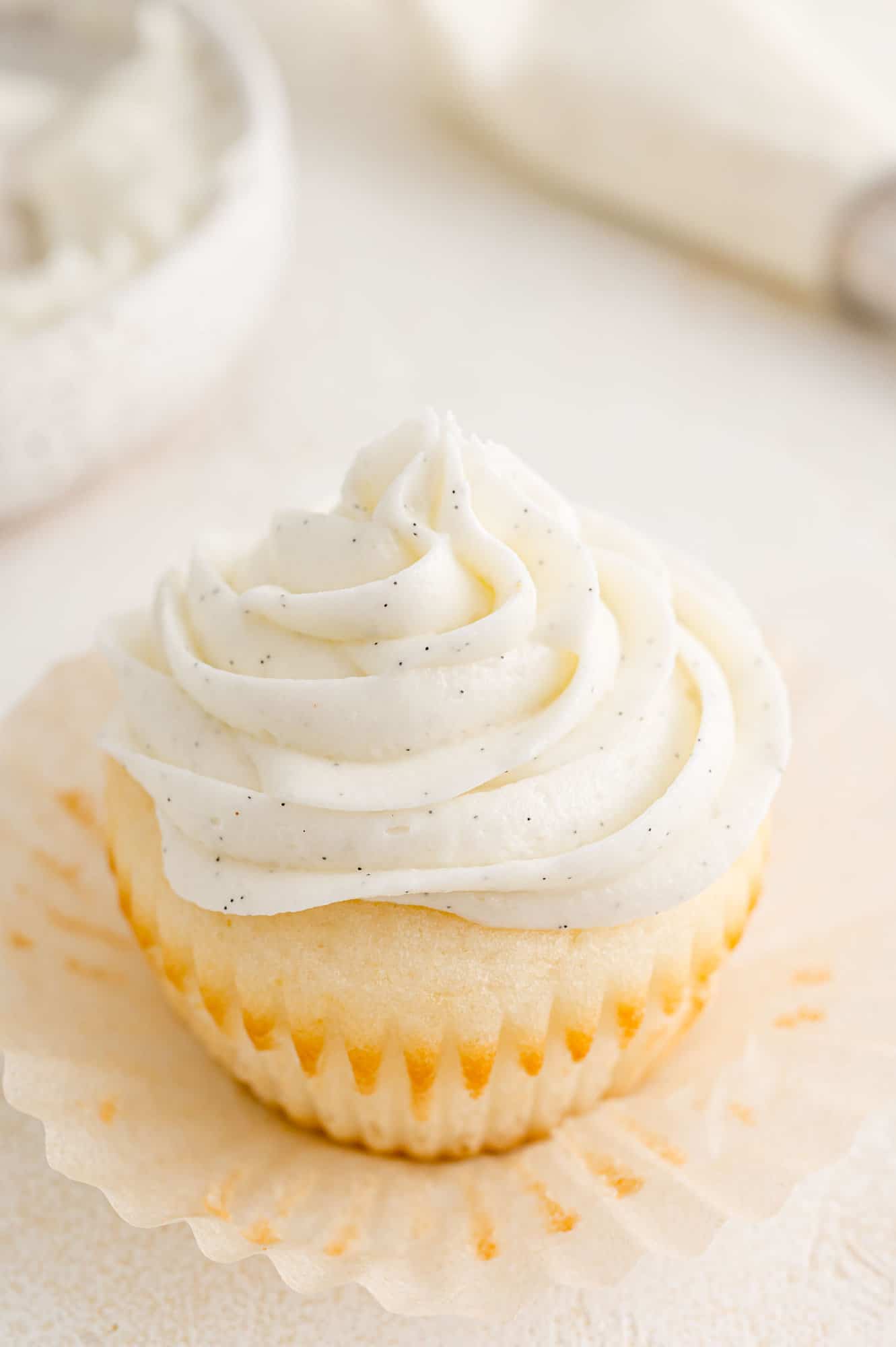 An unwrapped vanilla cupcake topped with a swirl of vanilla buttercream frosting, with a piping bag in the background.