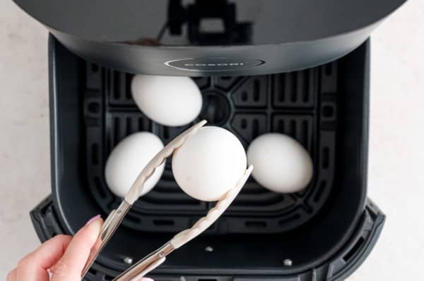 Tongs are used to lift boiled eggs from the basket of an open air fryer.