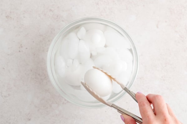 A hand uses tongs to drop a hard boiled egg into a bowl of ice water.