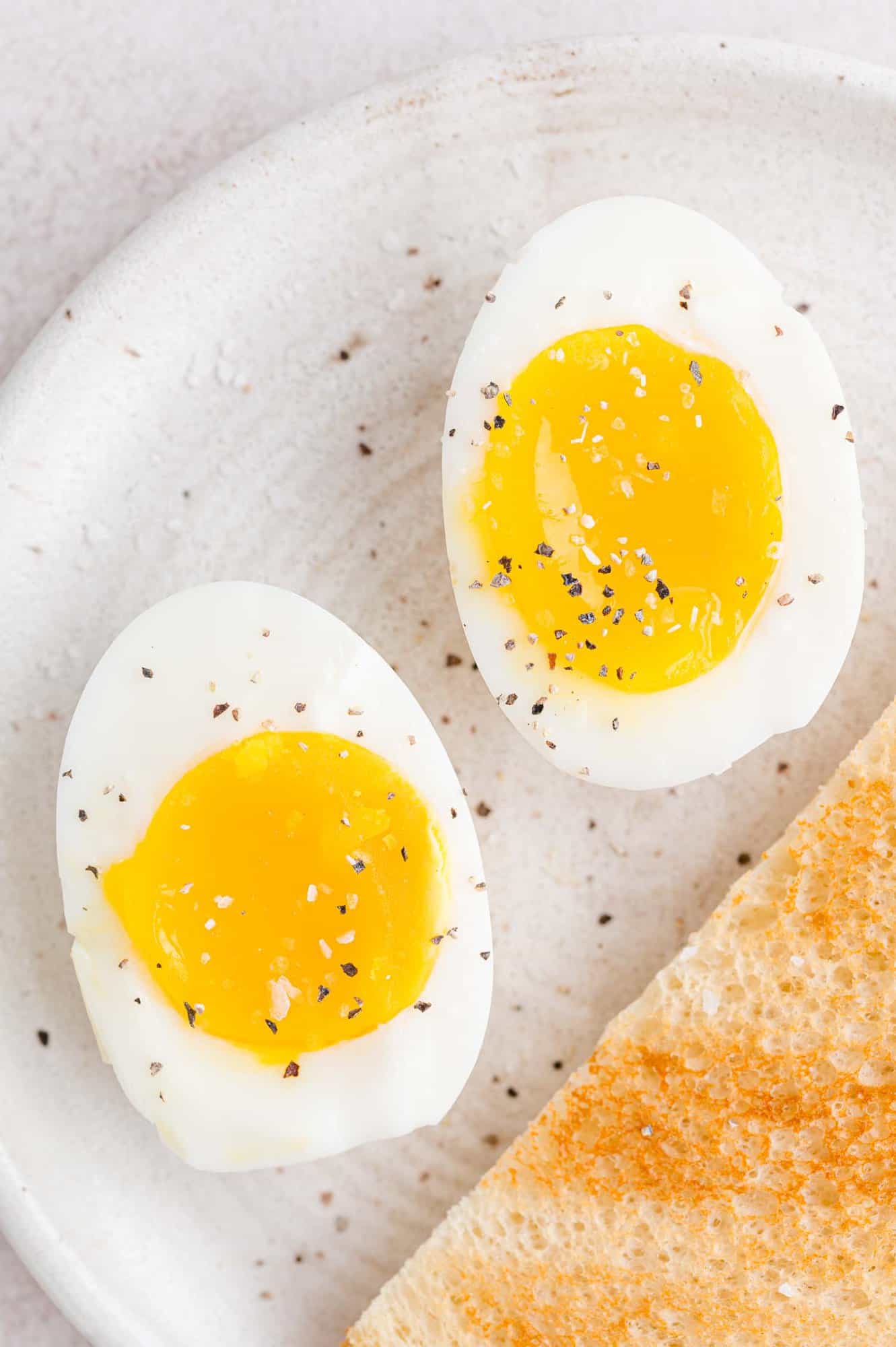 Close up of two halves of a boiled egg on a plate, seasoned with salt and pepper next to a slice of toast.