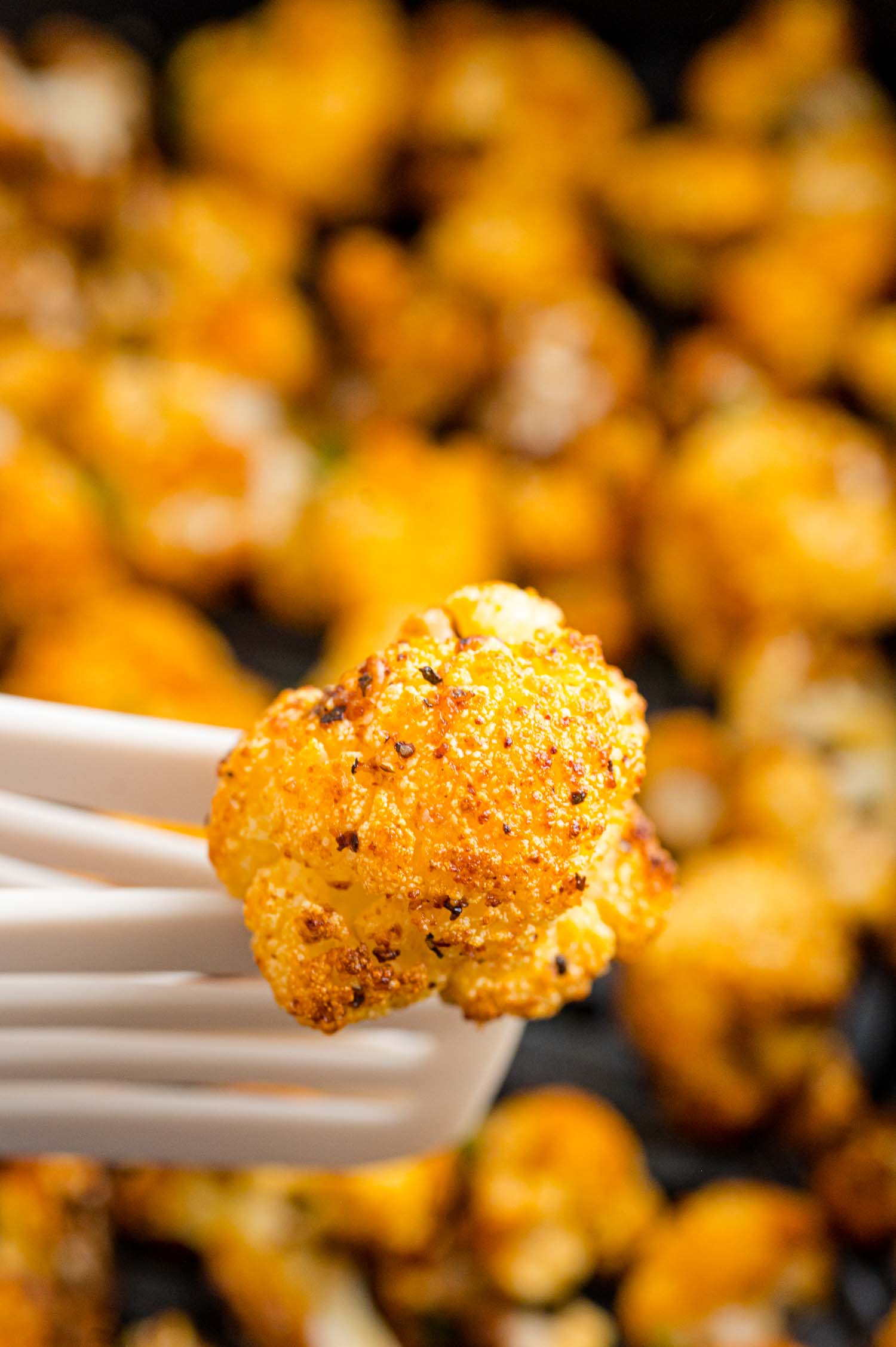 Close up of an air fried cauliflower floret lifted up on a spatula, with more cauliflower in the air fryer in the background.