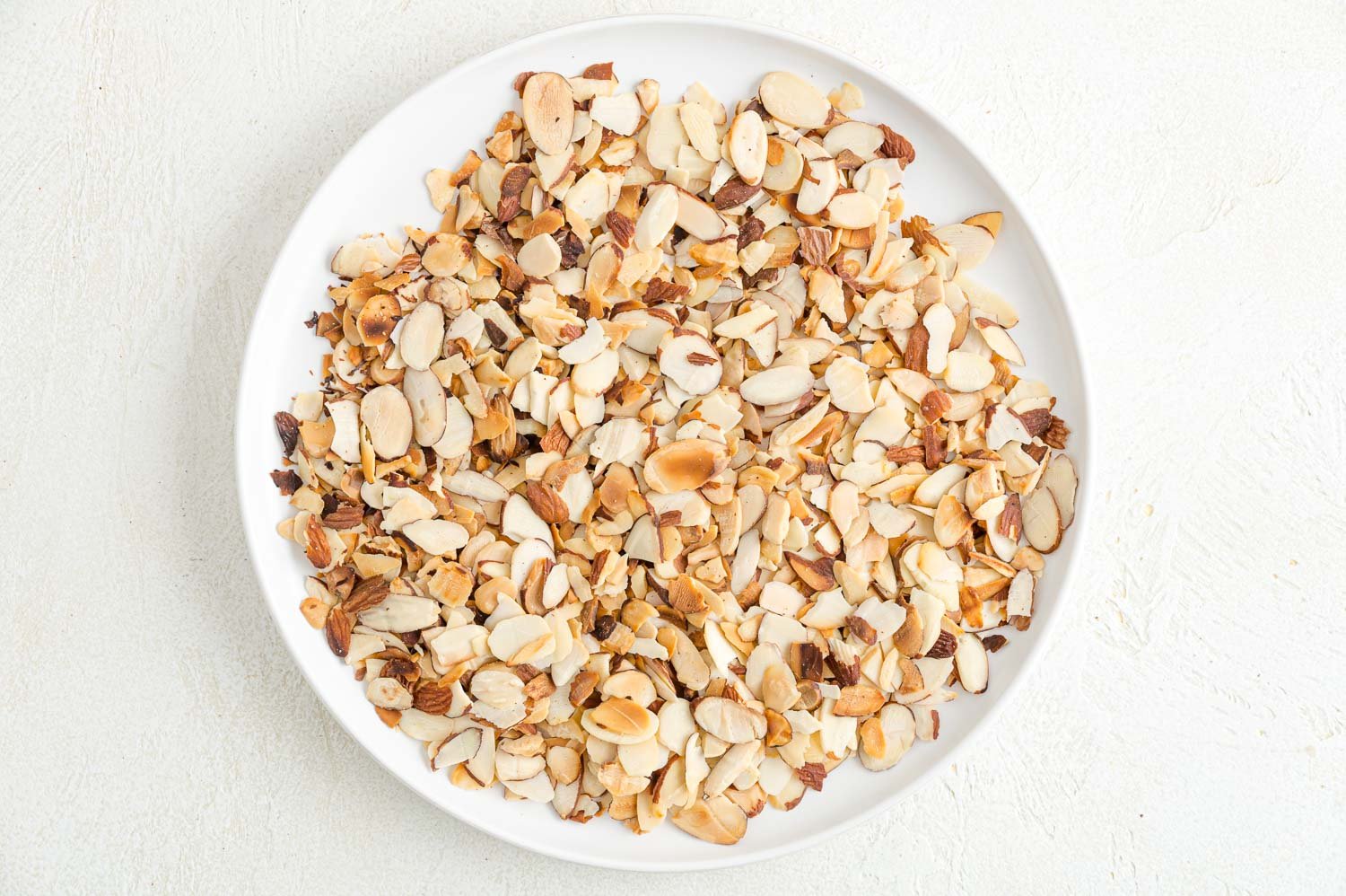 Toasting almonds is a great way to bring out their crunch and flavor. Learn how to toast almonds in the oven, microwave, and on the stovetop! Toasted almond slices on white plate.