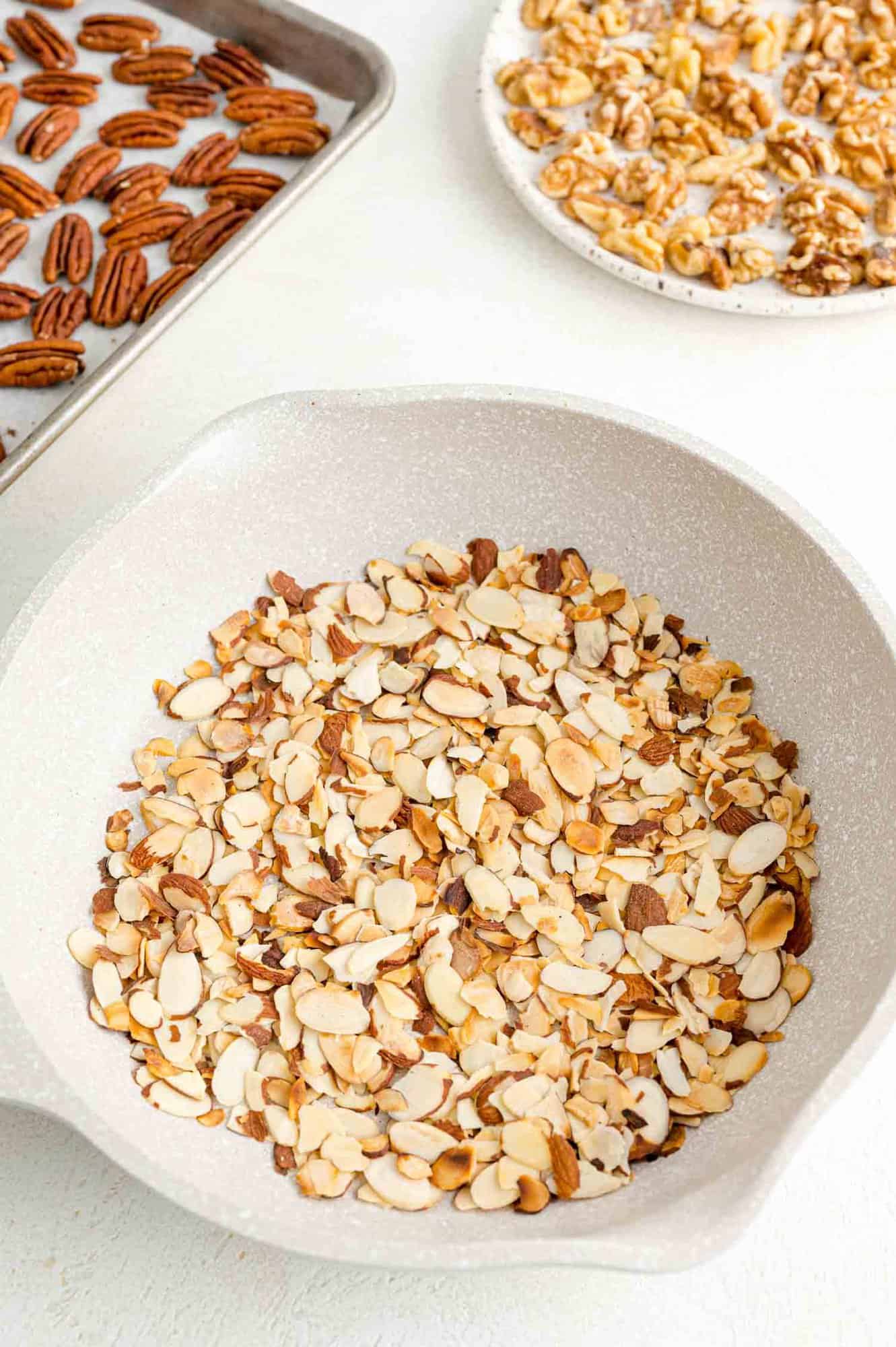 Toasted almonds in a white skillet next to a tray of toasted pecans and a plate of toasted walnuts.