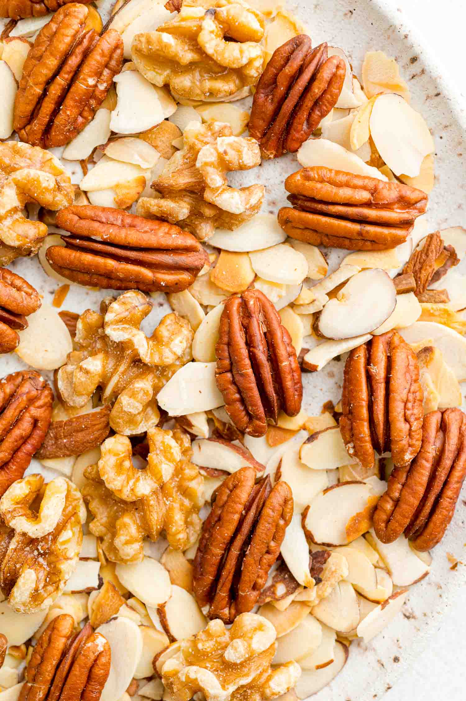 Close up of a medley of toasted almonds, walnuts, and pecans on a white plate.