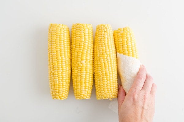 A hand uses a paper towel to pat corn on the cob dry after rinsing.