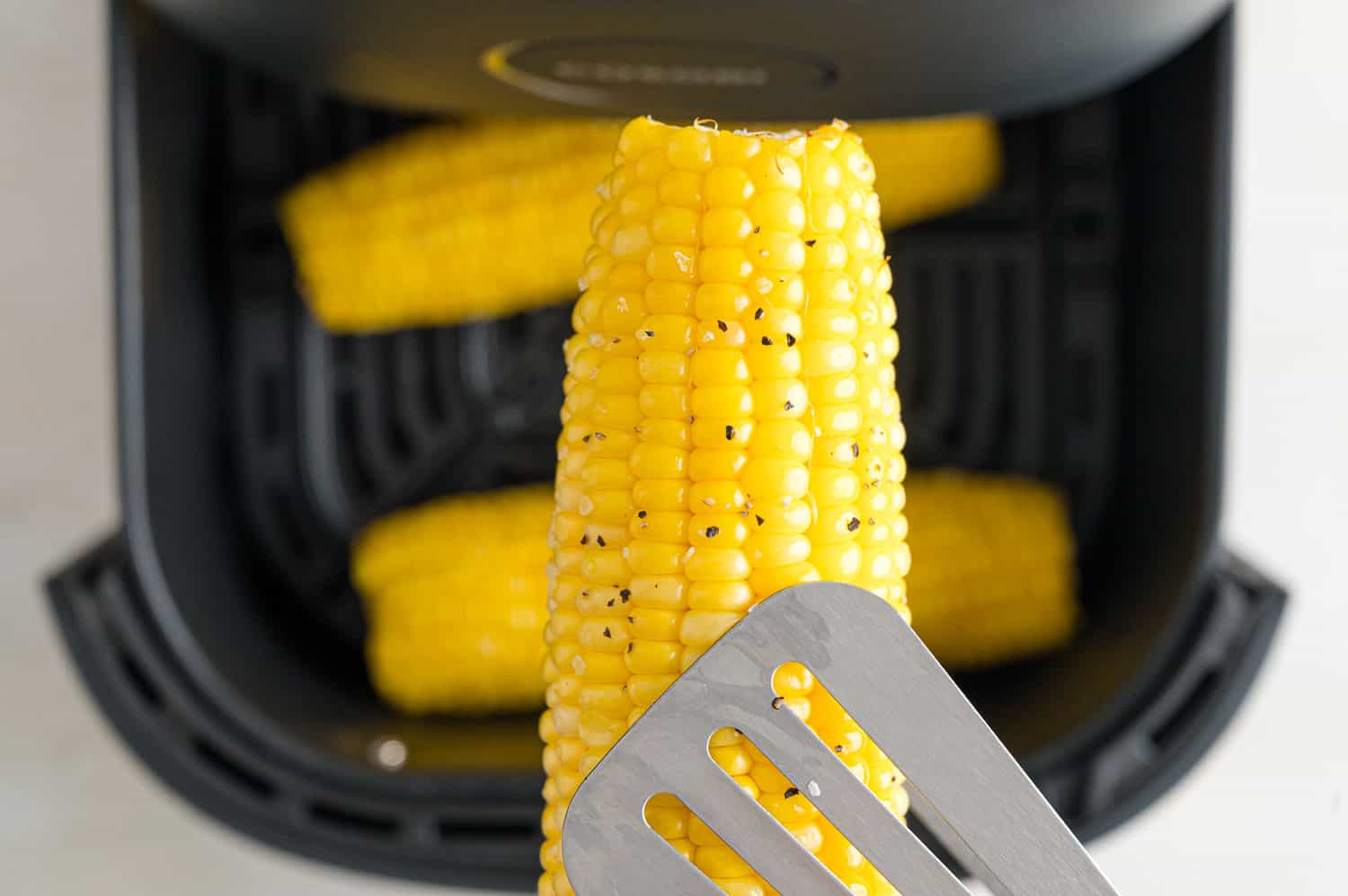 Close up of tongs holding an ear of corn above more corn inside the air fryer.