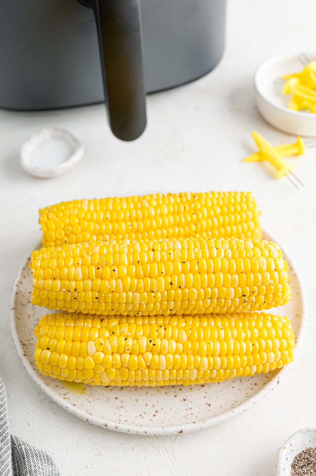 A stack of corn on the cob on a white plate next to the handle of an air fryer.