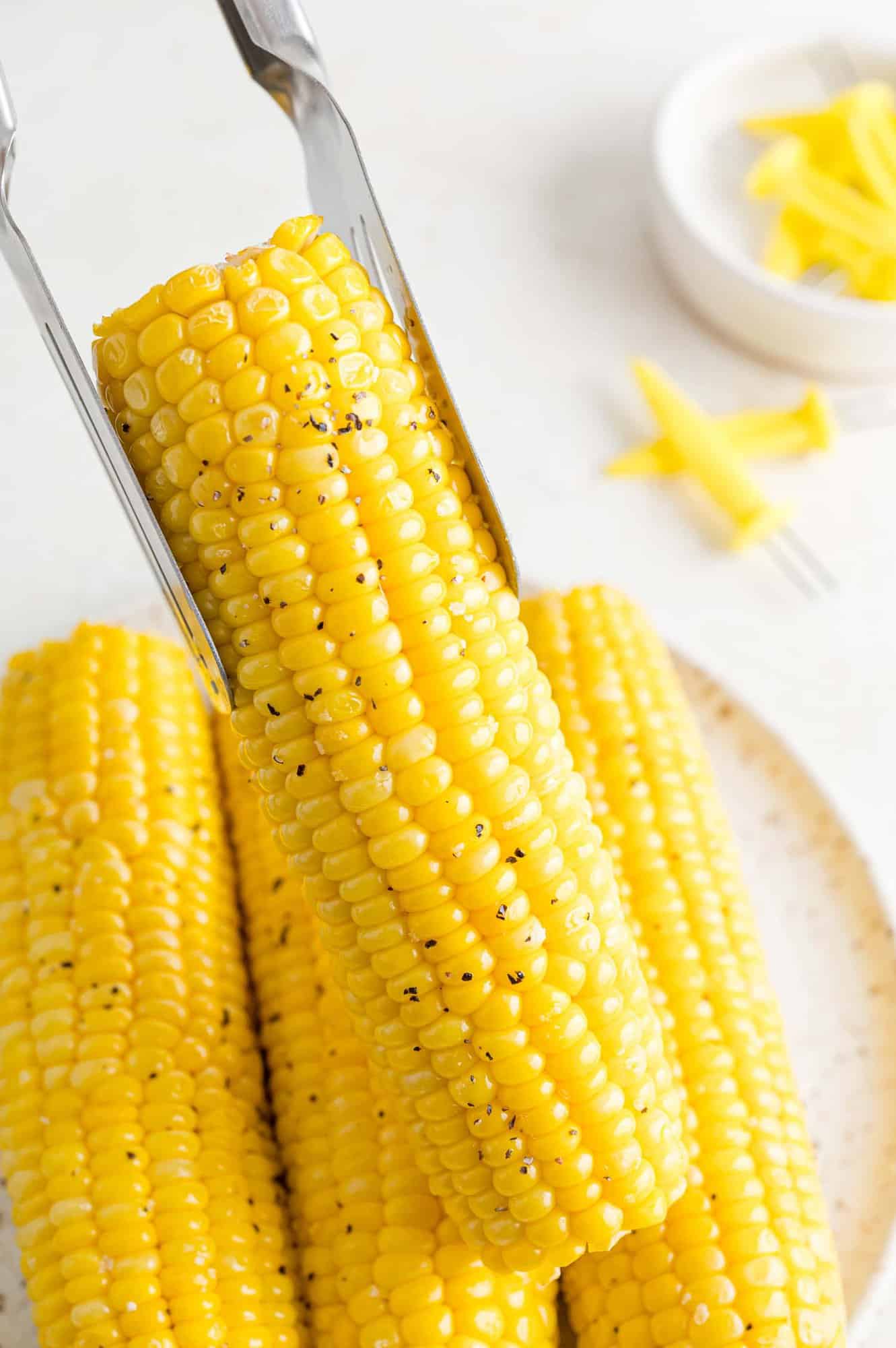 Tongs lifting an ear of corn from a plate with corn on the cob.