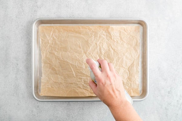 A hand holding a can of cooking spray over a baking sheet lined with brown parchment paper.