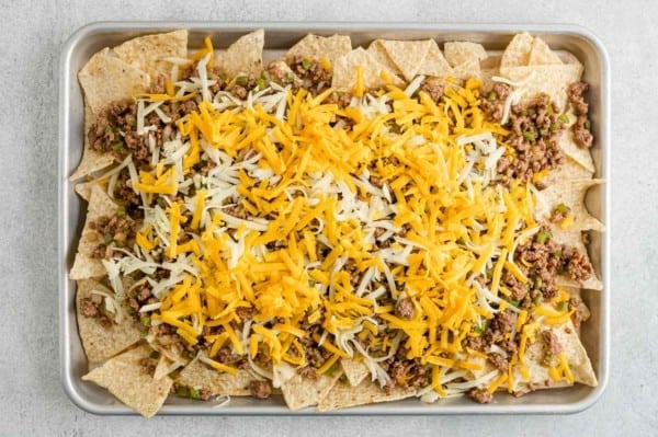 Tortilla chips topped with seasoned taco beef and shredded cheese on a parchment lined baking tray.