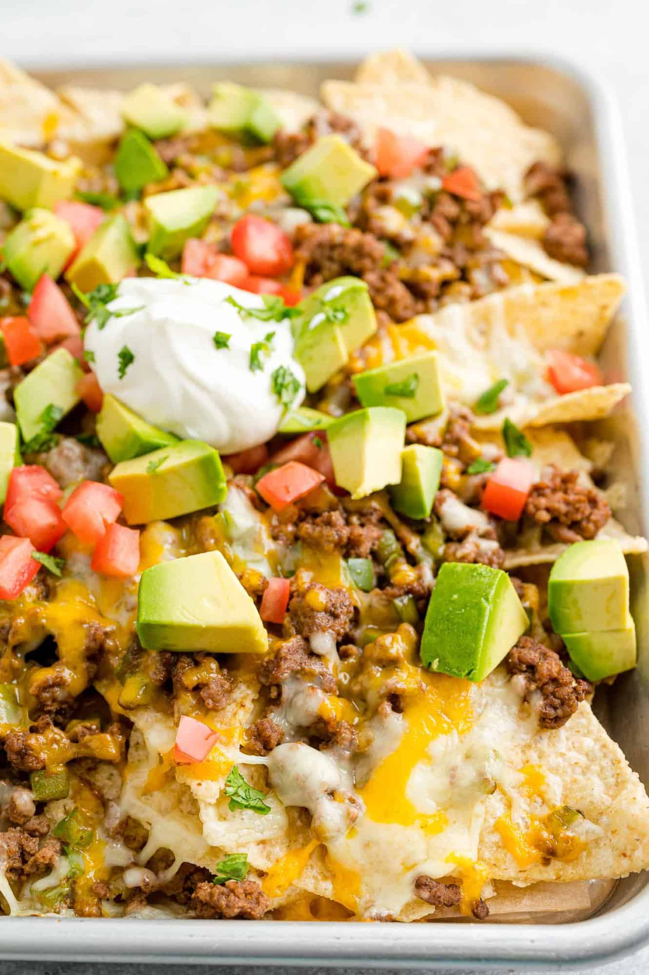 Close up view of a tray of beef nachos topped with cheese, diced tomatoes, avocados, onions, and dollops of sour cream.