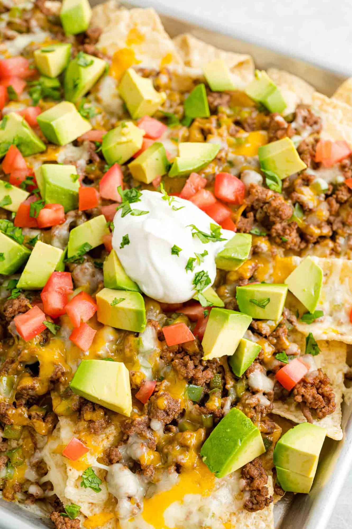 Close up view of a tray of beef nachos topped with cheese, diced tomatoes, avocados, onions, and dollops of sour cream.