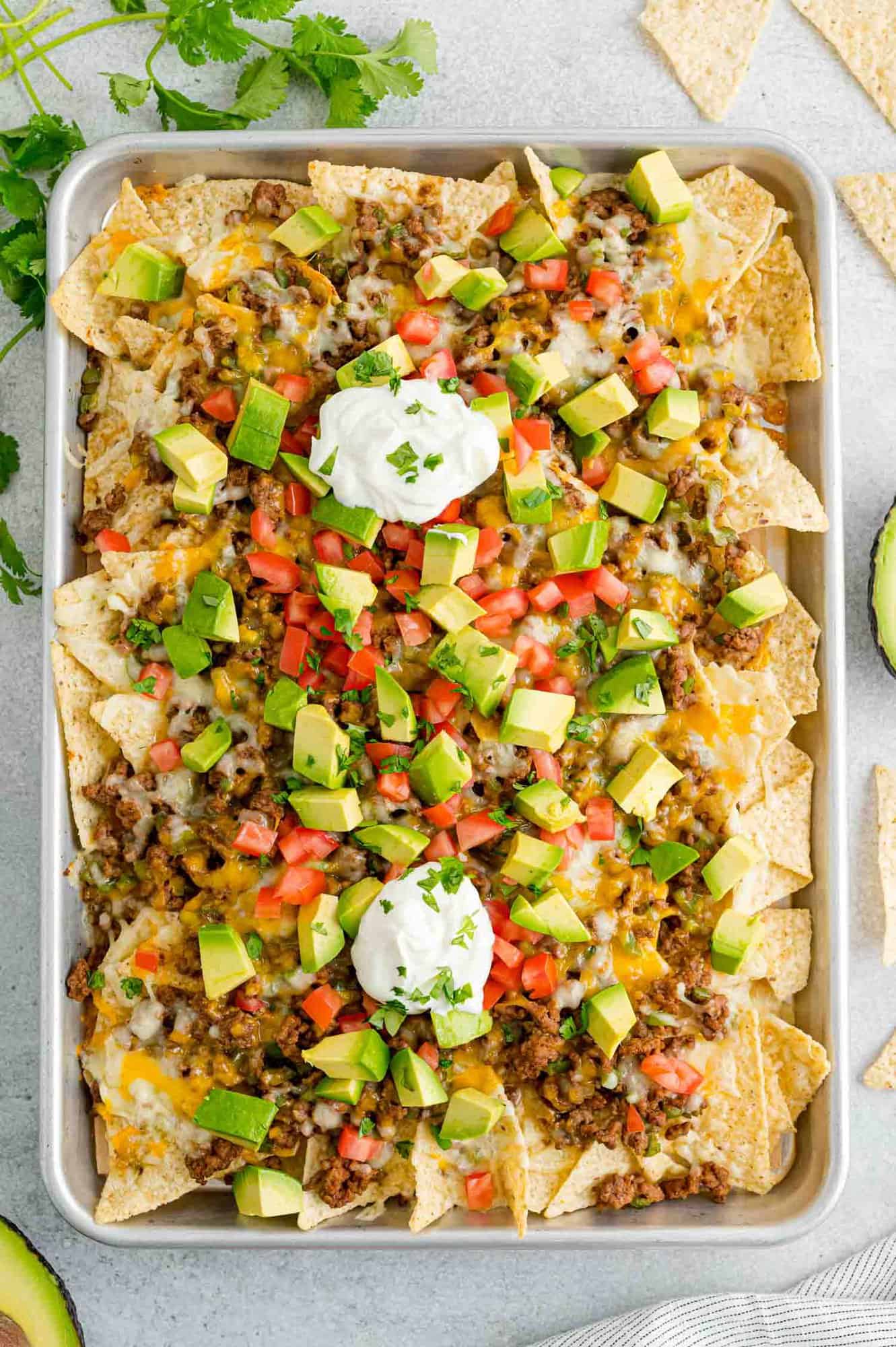 Overhead view of a tray of beef nachos topped with cheese, diced tomatoes, avocados, onions, and dollops of sour cream.