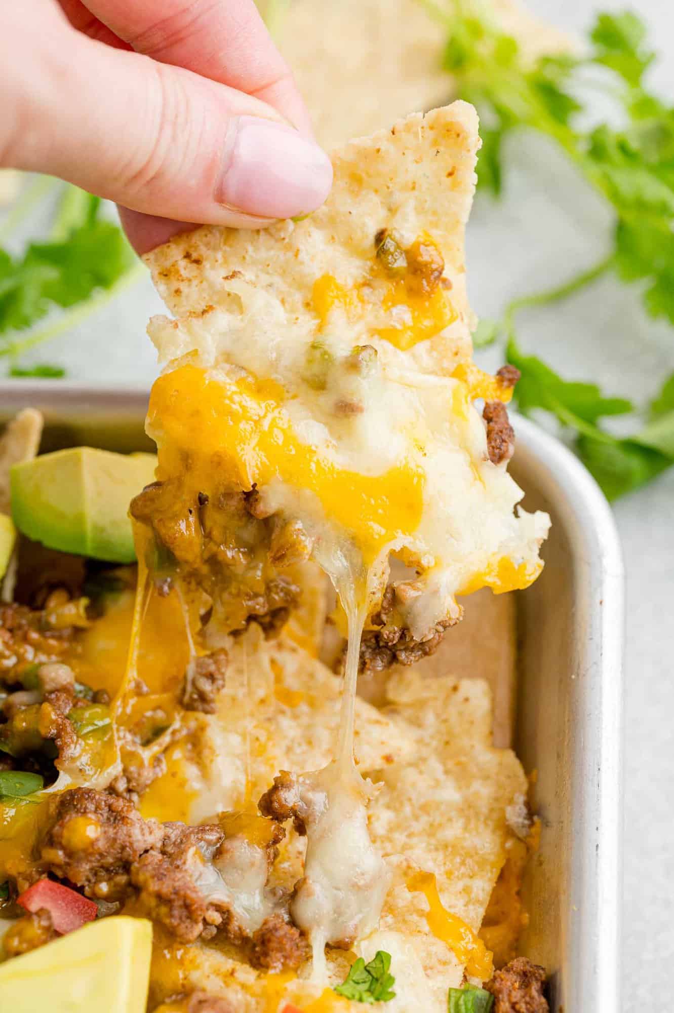 A hand lifts a cheesy tortilla chip from the corner of a pan of beef nachos.