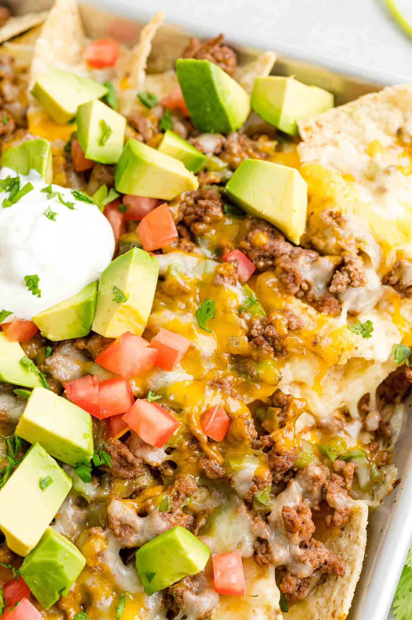 Close up view of a tray of beef nachos topped with cheese, diced tomatoes, avocados, onions, and dollops of sour cream.