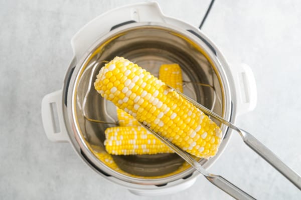 Overhead view of tongs holding an ear of corn above the open Instant Pot filled with more corn.