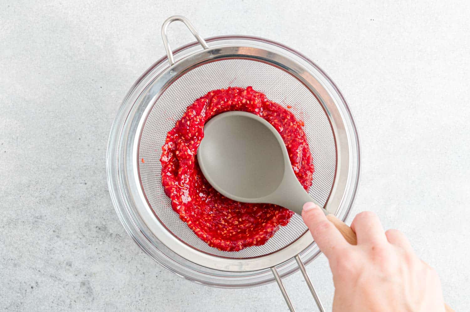 Spoon pressing raspberry puree through a fine mesh strainer.