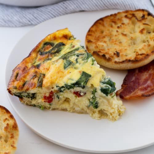 Slice of crustless quiche on a plate with english muffin and bacon.