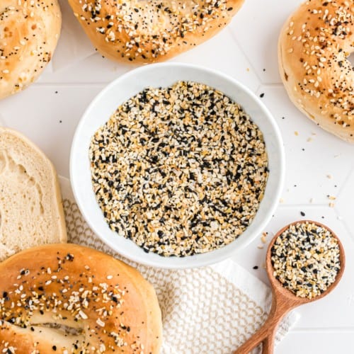 A wooden spoonful of everything bagel seasoning next to a bowl next to an everything bagel.