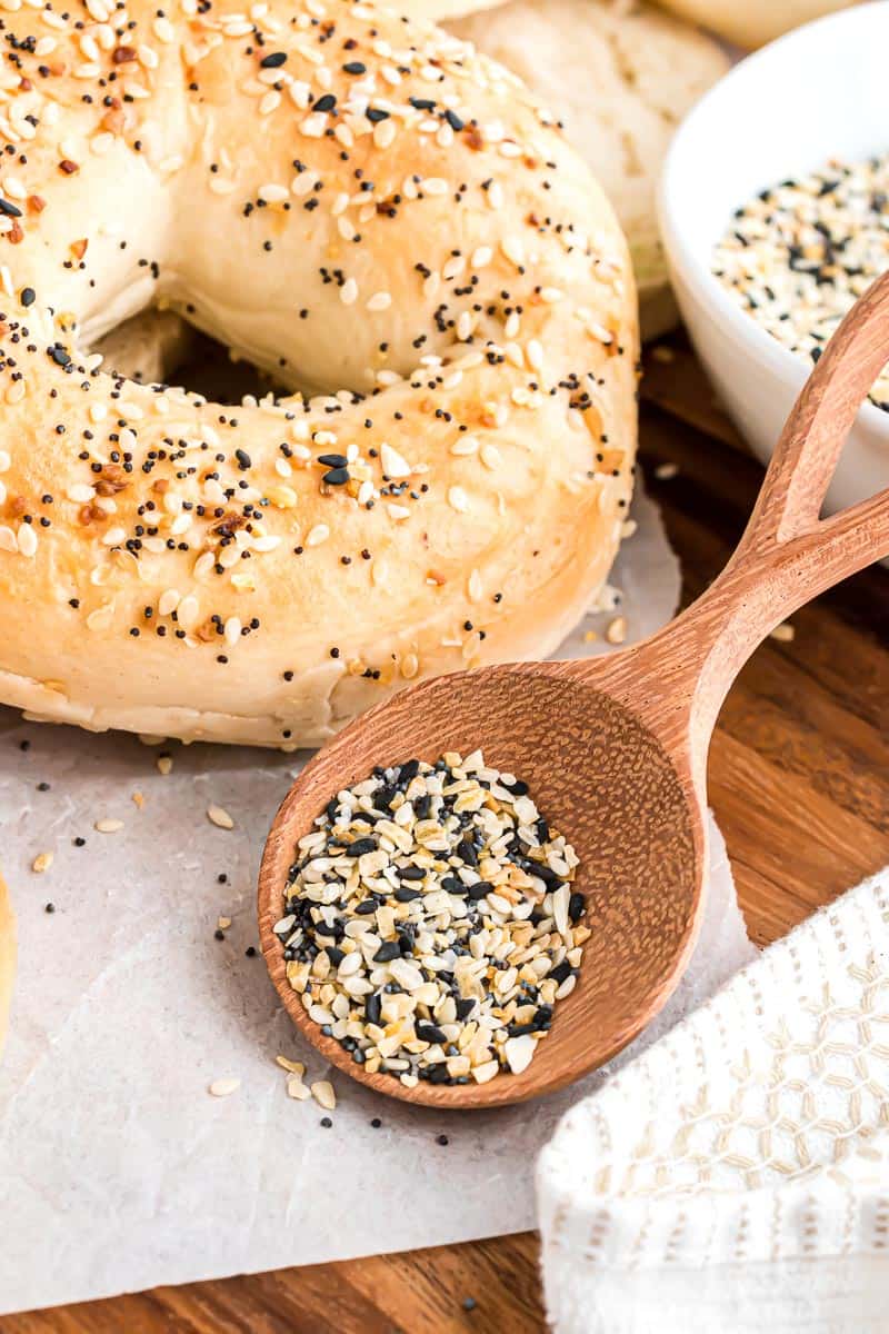 A wooden spoonful of everything bagel seasoning leaning against a bowl next to an everything bagel.