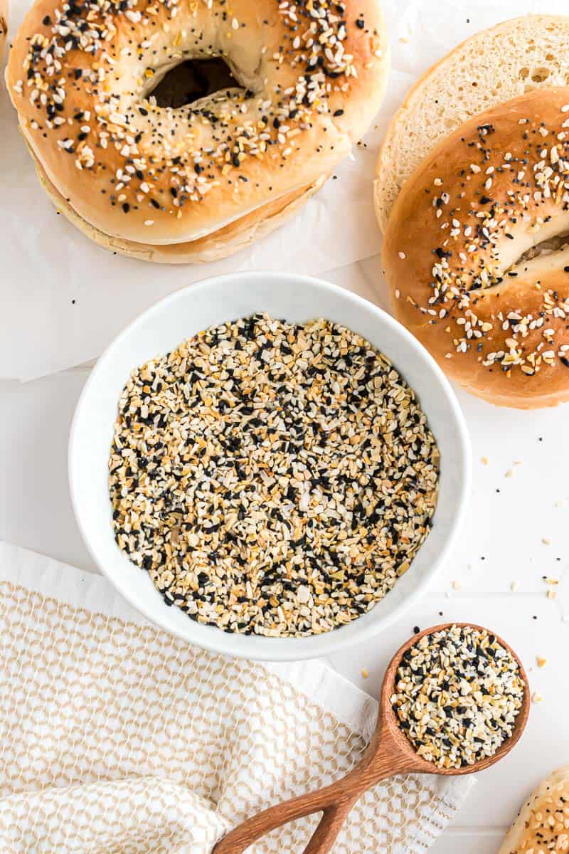 A bowl of everything seasoning next to a spoonful, and two everything bagels.