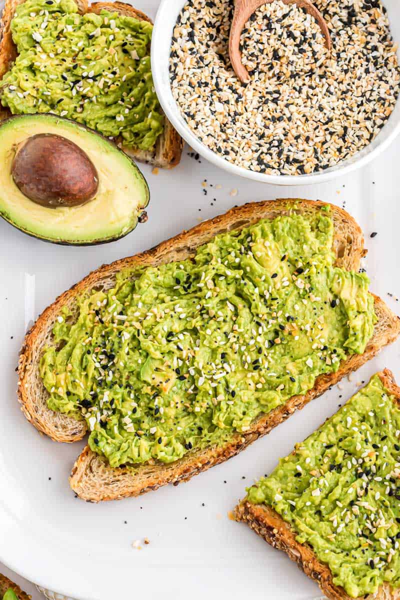 Overhead view of smashed avocado toast topped with everything bagel seasoning, next to a bowl of seasoning and half of an avocado.