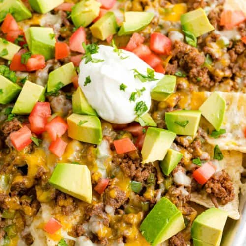 Close up view of a tray of beef nachos topped with cheese, diced tomatoes, avocados, onions, and dollops of sour cream.