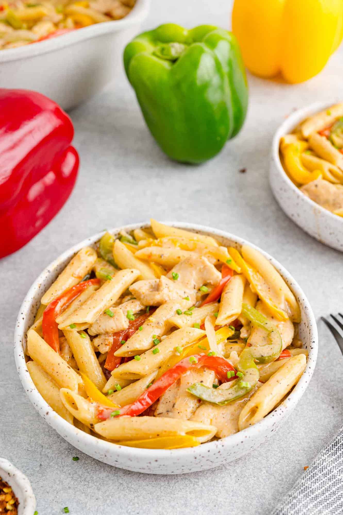 A bowl of Rasta Pasta next to red, green, and yellow bell peppers on a countertop.