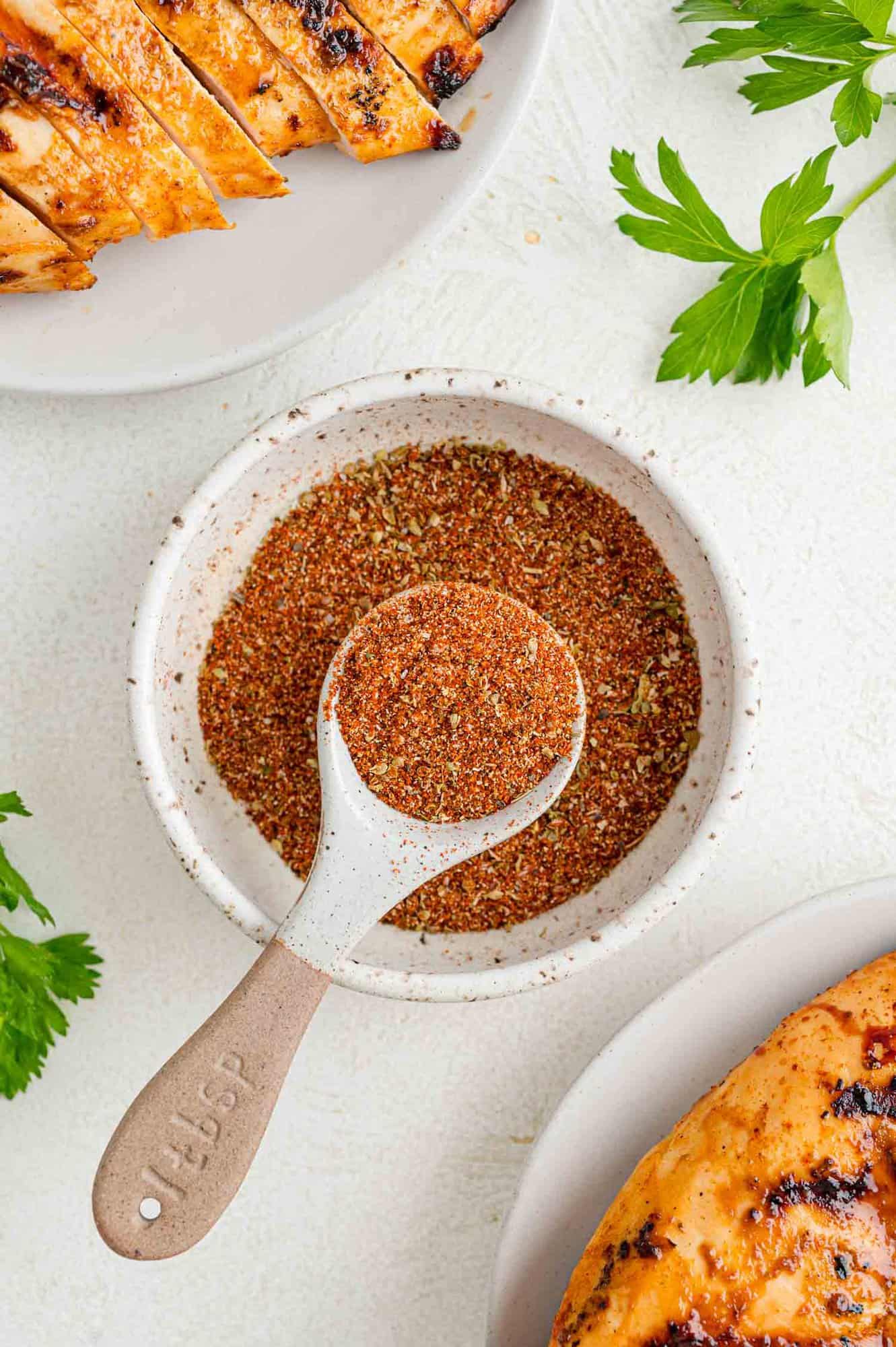 A bowl of Southwest spice blend in a bowl with a spoon, next to two plates with grilled chicken.