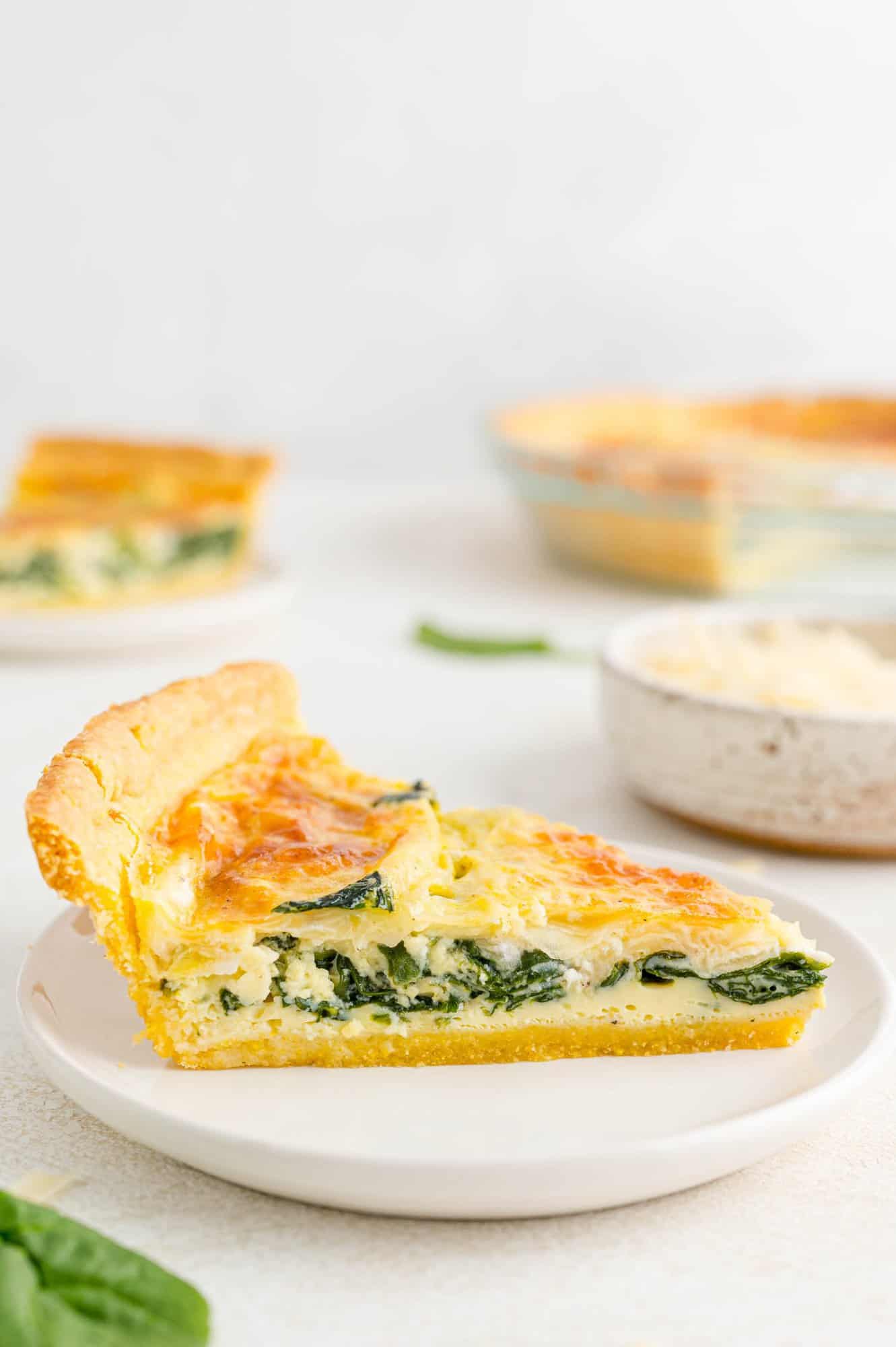 Spinach-Quiche-with-cornmeal-crust-slice-2 Slice of quiche on a white plate, more quiche in background.