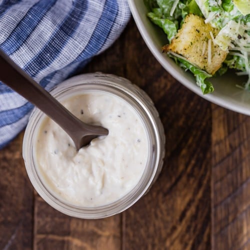 Overhead view of a jar of homemade caesar salad dressing, and a partially visible bowl of caesar salad.