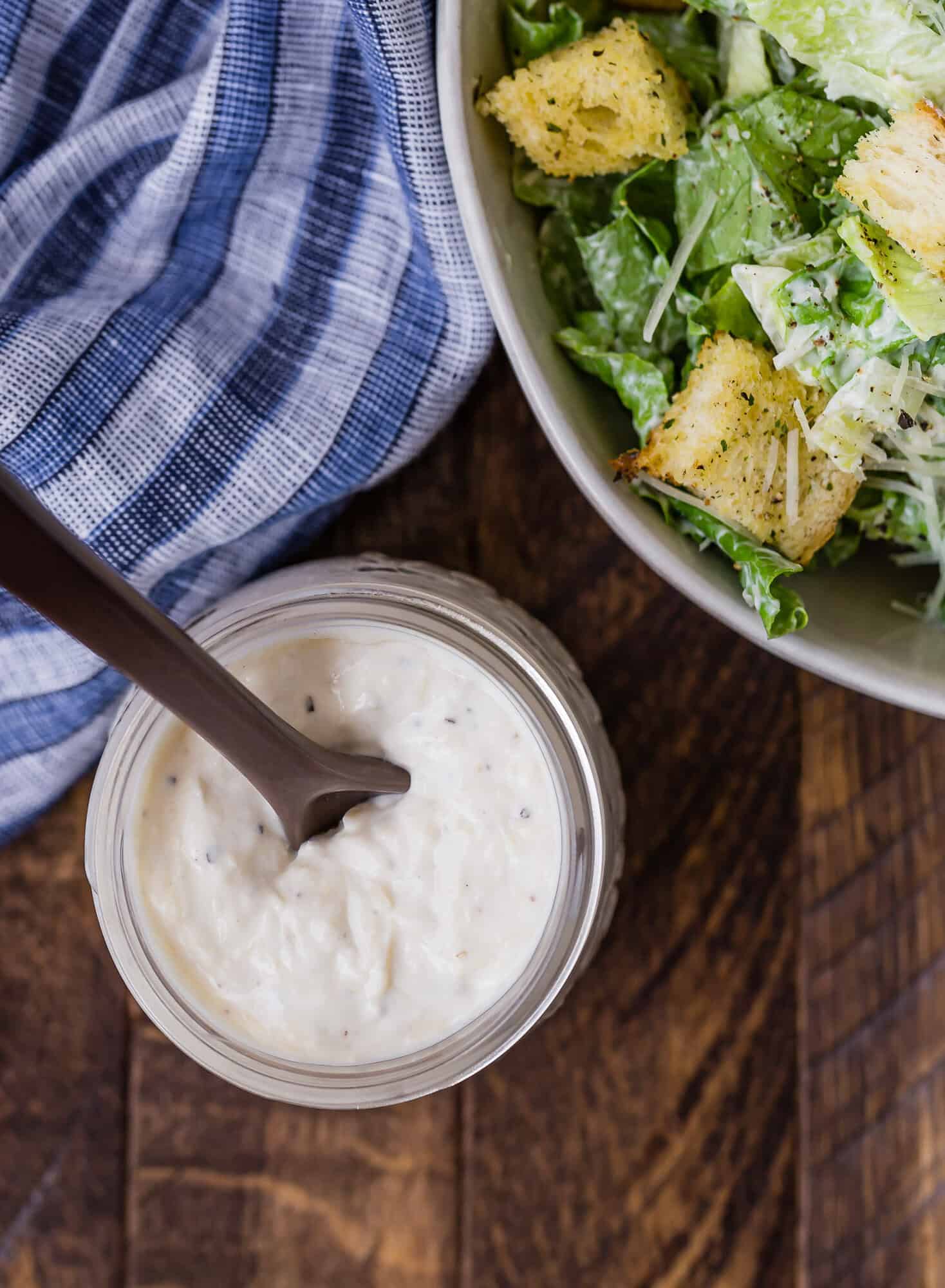 Overhead view of a jar of homemade caesar salad dressing, and a partially visible bowl of caesar salad.