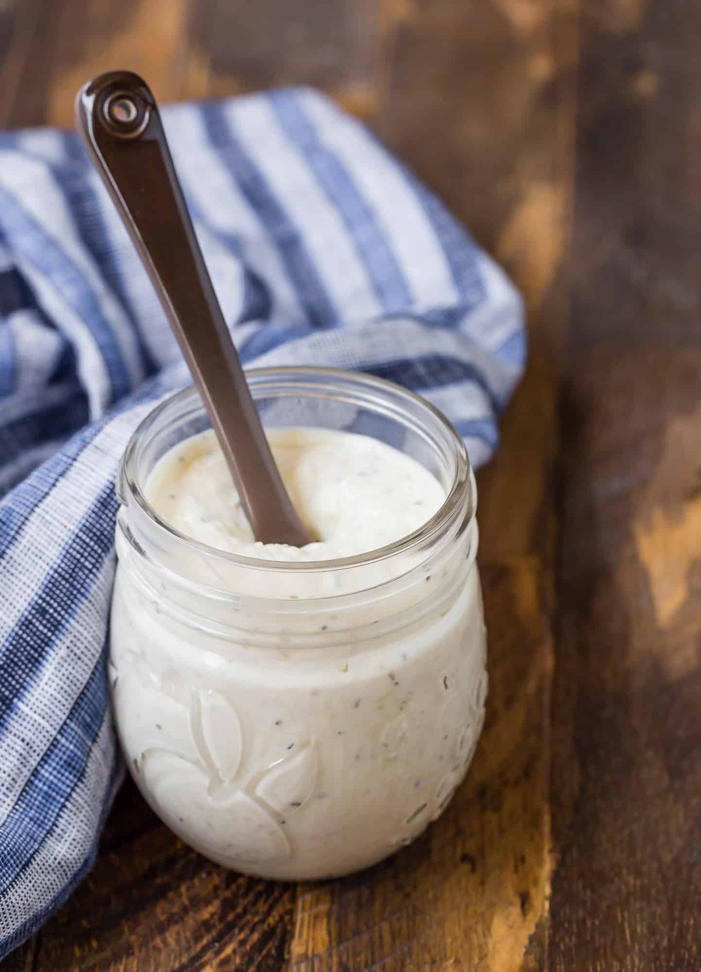 Small glass jar of a creamy caesar dressing on a wooden background. A blue and white linen is also pictured, and a small brown glass spoon.