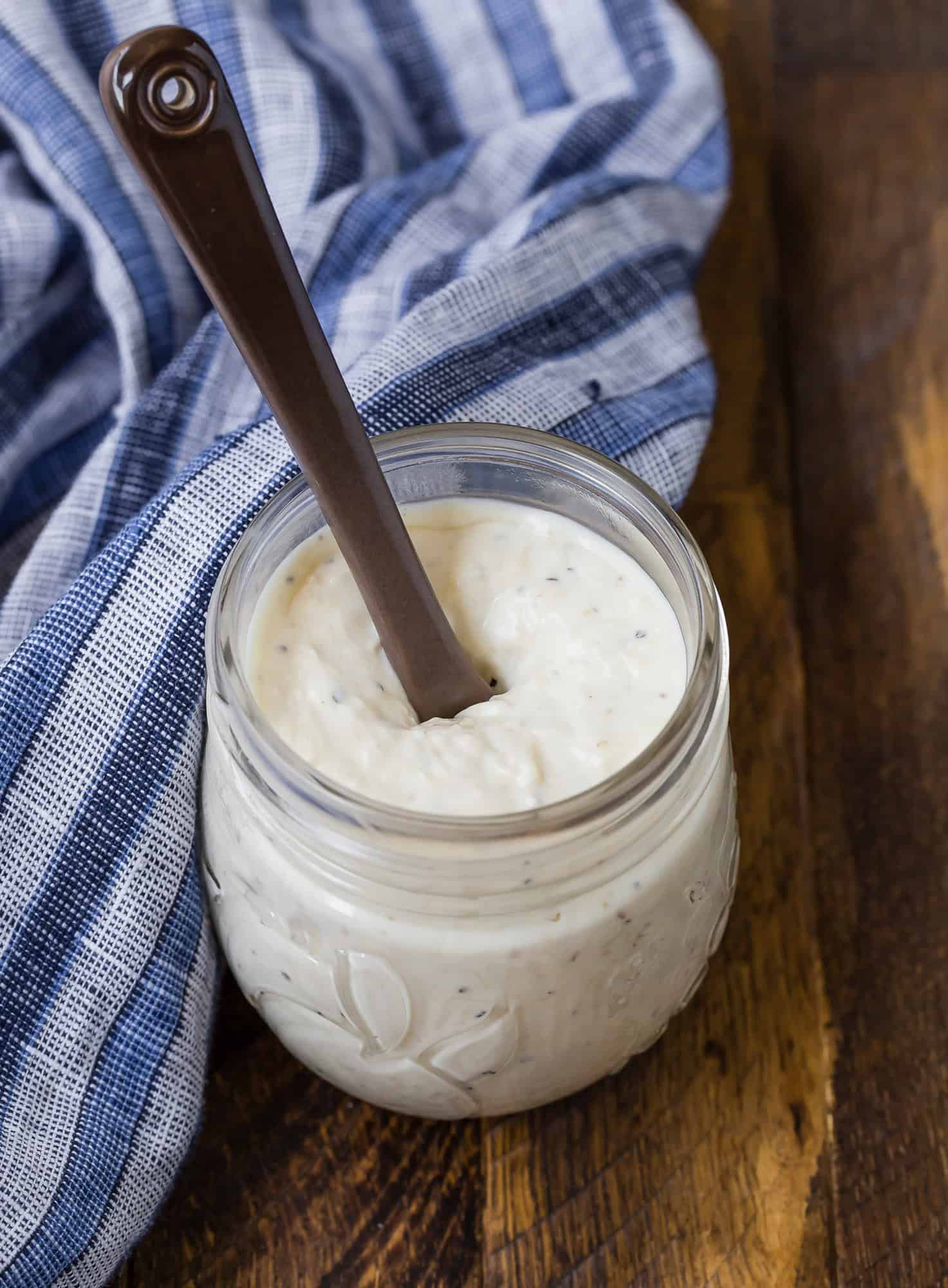 Small glass jar full of creamy dressing. Brown wooden background, blue and white linen.